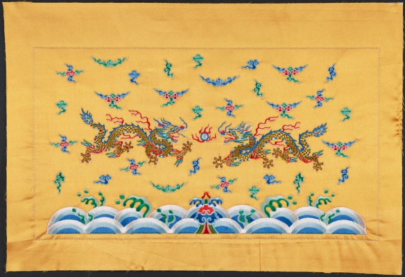 Une broderie dorée de deux dragons dont les griffes sont pointées vers une perle; ils volent parmi les nuages, au-dessus des vagues de l’océan.