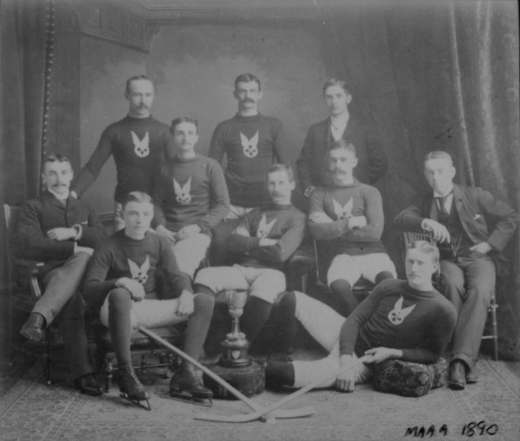 Photographie en noir et blanc montrant un groupe d’hommes debout, assis ou allongés. La plupart portent un uniforme d’équipe et des patins de hockey.