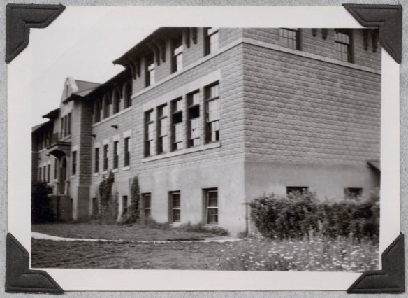 Photo noir et blanc légèrement floue, montrant le côté et la façade d’un bâtiment avec l’entrée principale.