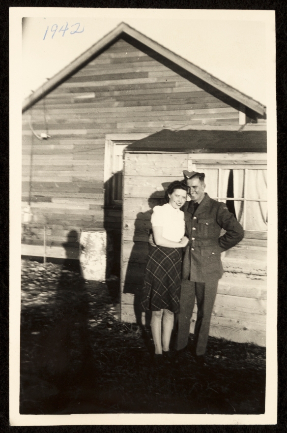 Photographie en noir et blanc d’un militaire en uniforme, le bras autour d’une jeune femme vêtue d’une robe fleurie, devant une maison de planches.