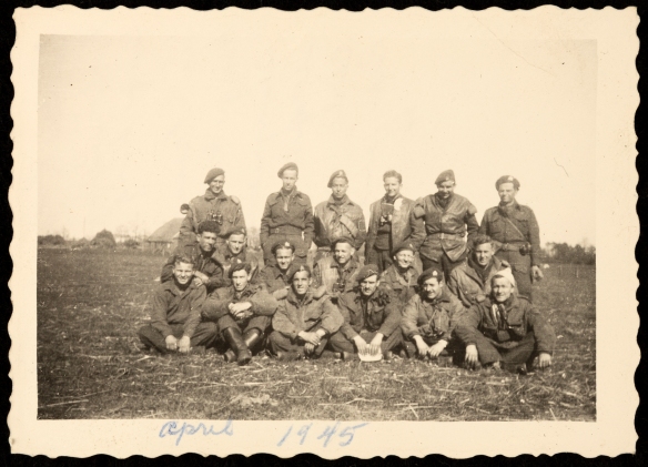 Photographie en noir et blanc d’un groupe de 18 soldats en uniforme, dans un champ cultivé.