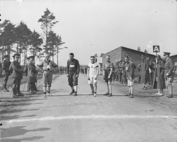 Une photographie noir et blanc de cinq hommes alignés sur une route, prêts pour le départ d’une course. Des soldats debout de chaque côté de la route les regardent.