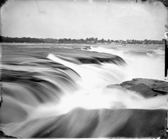 Photo noir et blanc d’un gros plan des chutes de la Chaudière avec des bâtiments visibles sur la rive éloignée.