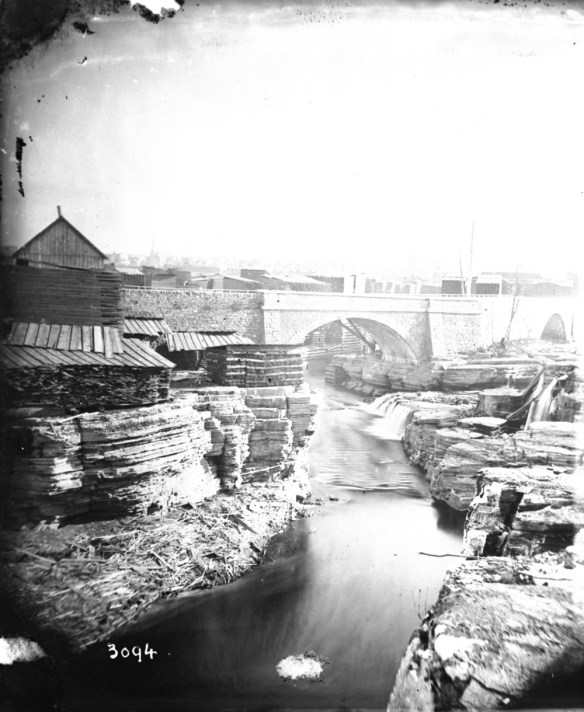Photo noir et blanc d’un paysage riverain industrialisé montrant un pont, des roches striées et des bâtiments en arrière-plan.