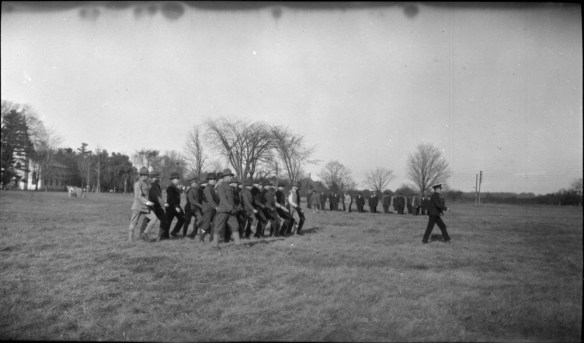 Photographie en noir et blanc d’un champ où un groupe d’officiers, à l’avant-plan, suivent leur commandant tandis que d’autres officiers en ligne sont au garde-à-vous, à l’arrière-plan.