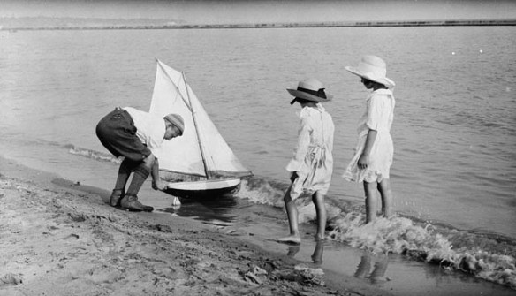 Une photographie noir et blanc de deux fillettes observant un garçon mettre à l’eau un voilier miniature. 