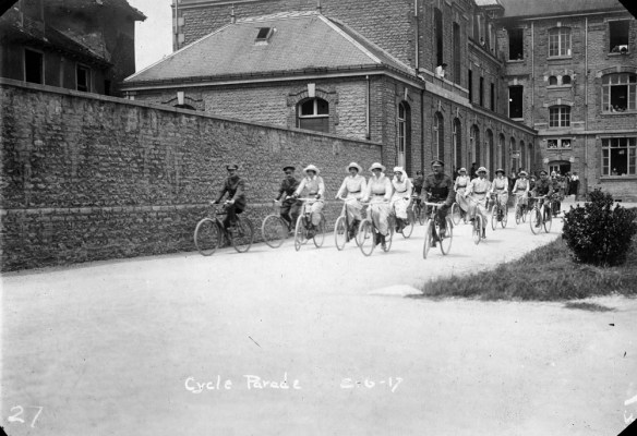 Photo noir et blanc d’hommes et de femmes en uniforme faisant du vélo. Les femmes sont vêtues d’uniformes de couleur pâle avec des ceintures foncées et des chapeaux alors que les hommes portent des uniformes militaires et des chapeaux. Ils pédalent le long d’une route bordée à gauche par un grand mur de briques. Un grand bâtiment aux façades fenêtrées est bien visible à l’arrière-plan. La légende « Cycle Parade » (Défilé à vélo) est inscrite au bas de l’image.