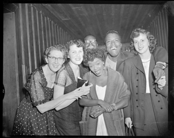 Photo noir et blanc de cinq jeunes personnes rassemblées autour de la chanteuse de jazz américaine Sarah Vaughan pour se faire photographier.