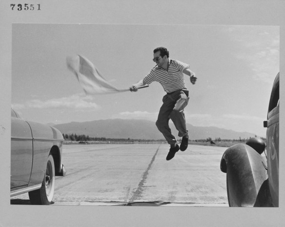 Une photographie noir et blanc d’un homme sautant dans les airs entre deux autos, agitant un drapeau pour donner le signal de départ d’une course. 