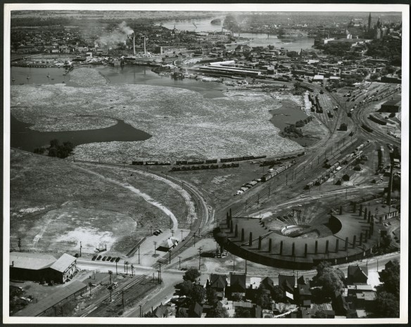 Photo aérienne noir et blanc d’un paysage industriel avec des rondins flottant dans l’eau et une centrale électrique et des lignes ferroviaires au premier plan. En arrière-plan se trouve la cité parlementaire.
