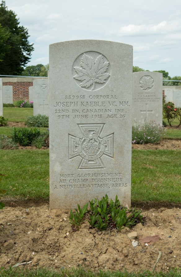 Photo couleur d’une pierre tombale devant laquelle quelques plantes commencent à pousser. On voit d’autres pierres tombales à l’arrière-plan.