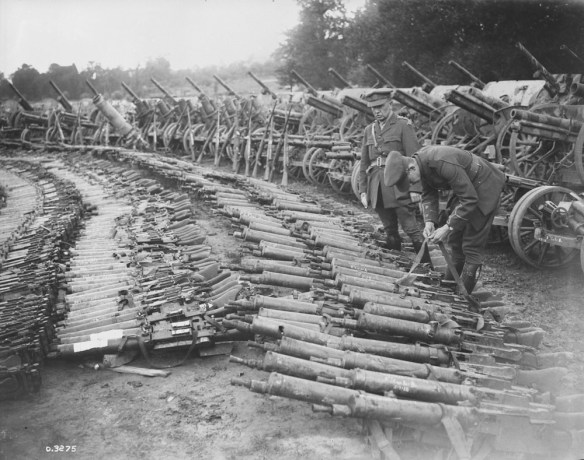 Photo noir et blanc de centaines de fusils et de mitrailleuses de diverses tailles reposant sur le sol. Un soldat, debout, regarde les armes tandis qu’un autre soldat se penche pour examiner la bandoulière d’un fusil.