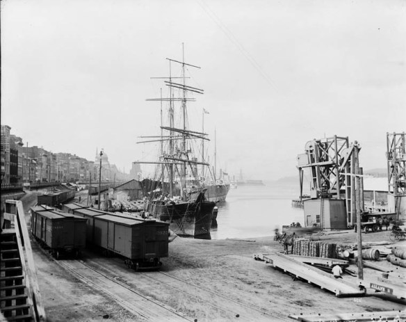 Une photographie en noir et blanc d’un chemin de fer longeant un quai où sont amarrés des bateaux.