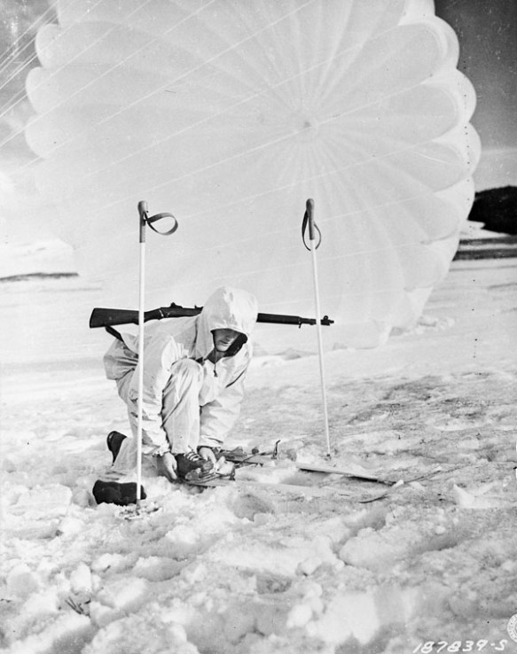 Photo noir et blanc d’un soldat portant une combinaison d’hiver blanche, accroupi pour ajuster ses fixations de ski. Un parachute est déployé derrière lui. 