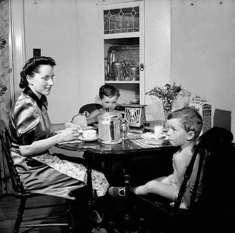 Photographie en noir et blanc d’une femme et de ses deux jeunes fils assis à une table en train de déjeuner.
