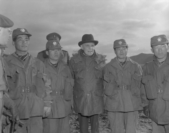 Photo noir et blanc de Louis St-Laurent entouré de militaires. Deux soldats sud-coréens se tiennent à sa gauche, et deux autres à sa droite. À l’arrière-plan, on distingue deux soldats canadiens. Au premier plan, à gauche, on aperçoit le profil partiel d’un soldat canadien portant des lunettes et un képi.
