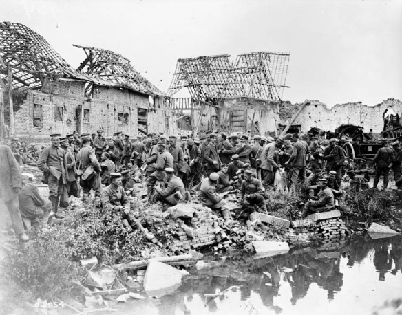 Photo noir et blanc montrant un large groupe de soldats allemands rassemblés entre un village et une rivière ou un canal. Les bâtiments à l’arrière-plan sont en bonne partie détruits.