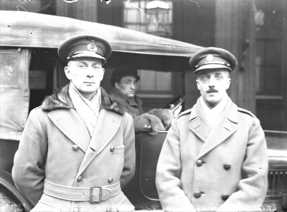 Photo noir et blanc de deux officiers devant une voiture dans laquelle le conducteur est assis. Ils portent tous deux une casquette d’officier et un pardessus.