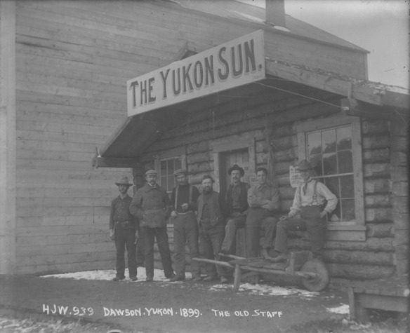 Photo noir et blanc d’un groupe d’hommes devant une cabane en bois rond dont l’enseigne indique « The Yukon Sun ».