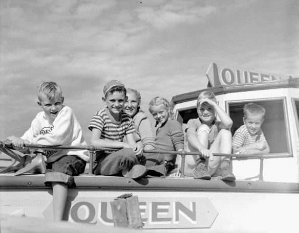 Photo noir et blanc de deux fillettes et de quatre garçons assis sur le pont avant du bateau à moteur Queen