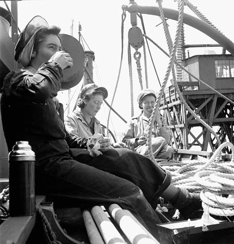 Photo noir et blanc de trois femmes prenant leur dîner dans une barque à côté de rames et d’une corde enroulée.