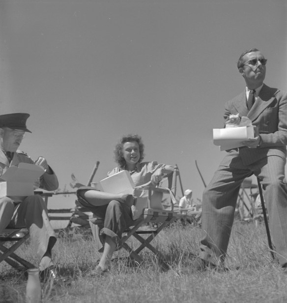 Photo noir et blanc de l’actrice Lucia Carroll et de deux membres de la distribution du film Les Chevaliers du ciel en train de dîner en plein air sur le lieu du tournage.