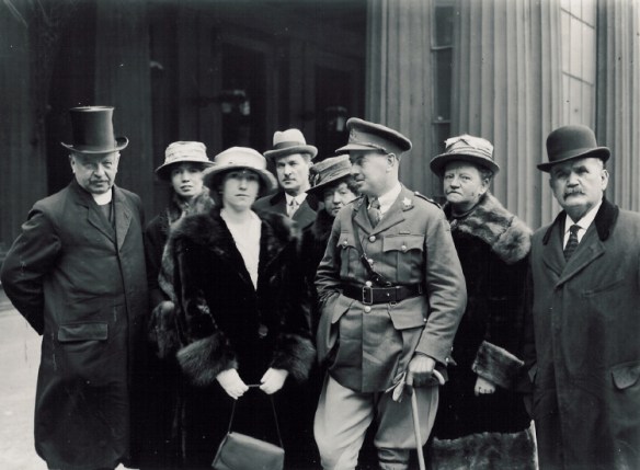 Photo noir et blanc de quatre femmes et de quatre hommes vêtus de manteaux chauds et coiffés de chapeaux devant le palais de Buckingham. L’homme au centre porte un uniforme militaire et s’appuie sur une canne.
