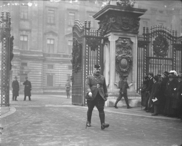Photo noir et blanc d’un homme moustachu vêtu d’un pantalon écossais, d’une ceinture Sam Browne et d’une casquette, passant des grilles ornementales avec un bâton de marche. À sa droite, une foule regarde vers les grilles.