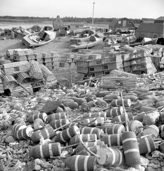 Photo noir et blanc d’un village côtier. On voit des homardiers à l’arrière-plan, des casiers à homard au centre, et des balises flottantes au premier plan.