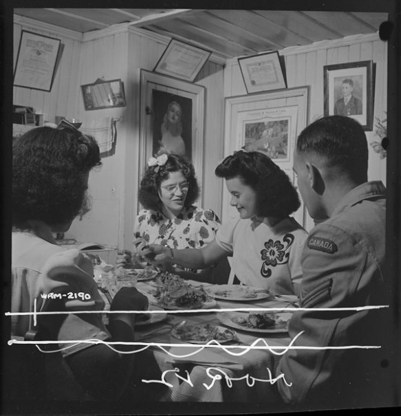 Photo noir et blanc de trois femmes et d’un homme partageant un souper dans la salle à manger d’une maison.