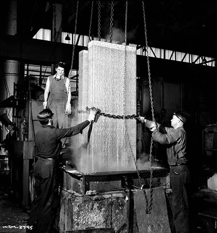 Une photographie noir et blanc d’une femme et de deux hommes manipulant et retirant des blocs d’aluminium de leurs moules à l’aide de chaînes.