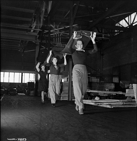 Une photographie noir et blanc de trois femmes transportant ensemble au-dessus de leur tête une longue feuille d’aluminium vers la table d’inspection.