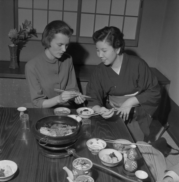 Photo noir et blanc de deux femmes dans un restaurant japonais, assises à une table couverte d’une variété de mets. La femme de droite explique à celle de gauche comment se servir de ses baguettes.