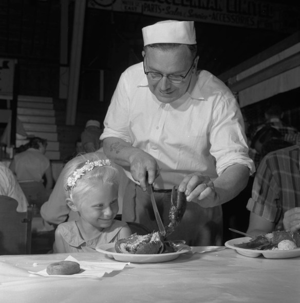 Photo noir et blanc d’un homme aidant une fillette attablée devant son repas de homard.