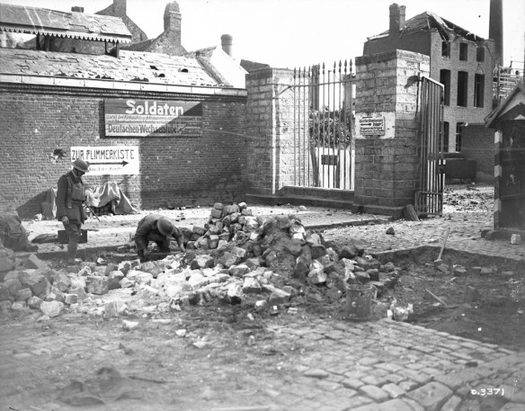 Photo noir et blanc d’un village où des bâtiments ont été endommagés. Au centre, on voit un imposant amas de pierres et de débris. Un soldat est agenouillé au sol, tout près. À gauche, un autre soldat se tient debout et l’observe.