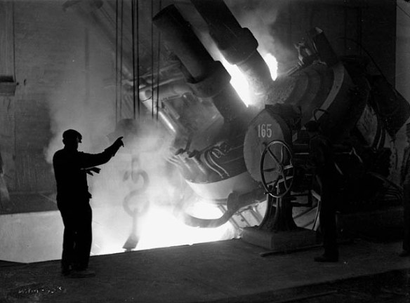 Photographie en noir et blanc d’un ouvrier, debout à côté d’un four, qui dirige le coulage d’acier en fusion dans une poche de coulée.
