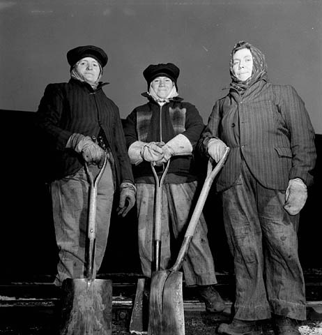 Photographie en noir et blanc de trois ouvrières du rail, portant des gants et de lourds vêtements de travail, et posant avec leurs pelles.