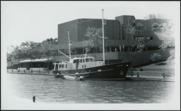 Photo noir et blanc d’un bateau de plaisance amarré le long d’un canal à proximité d’un imposant édifice. 