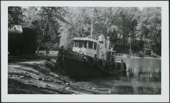 Photo noir et blanc d’un petit bateau-pompe amarré sur la rive boisée d’un cours d’eau. 