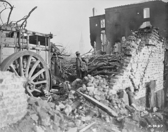 Photo noir et blanc d’un soldat en uniforme, dos à l’appareil photo, se tenant au milieu des décombres devant un bâtiment détruit. On aperçoit à gauche une charrette avec une grosse roue.