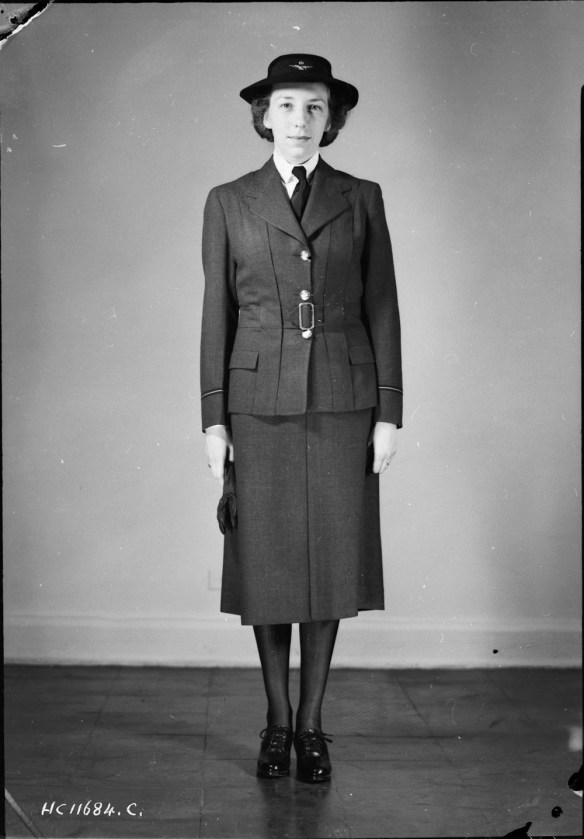 Photo noir et blanc d’une femme se tenant droite, vêtue d’ un uniforme militaire composé d’une veste Norfolk, d’une jupe et de chaussures noires.