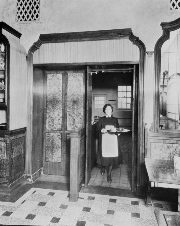 Photographie noir et blanc d’une femme sortant de la cuisine d’un restaurant en portant un plateau avec une théière et des tasses.
