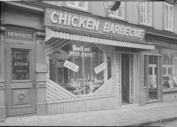 Photographie noir et blanc de l’extérieur du restaurant Nick's Chicken Barbecue. Une enseigne au néon dans la fenêtre annonce « Good Food » [bonne nourriture] et « Beer & Wine » [bière et vin].