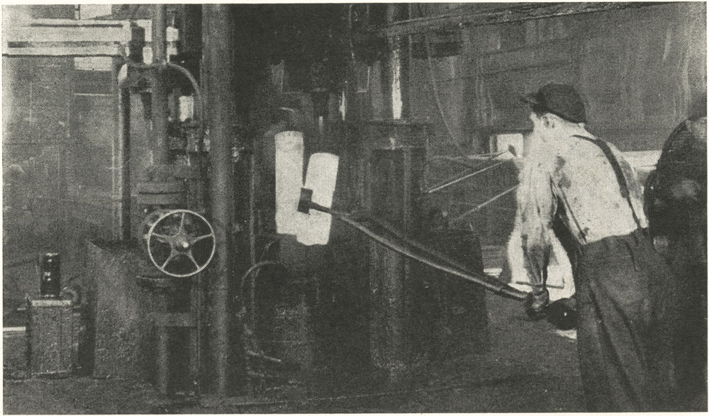 Photographie noir et blanc montrant un ouvrier manipulant un lingot d’acier brûlant à l’aide de longues pinces.