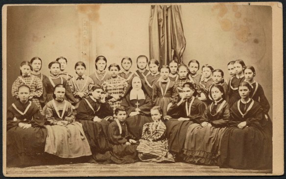 Portrait noir et blanc de 27 jeunes filles, médaille au cou, assises autour d’une religieuse.