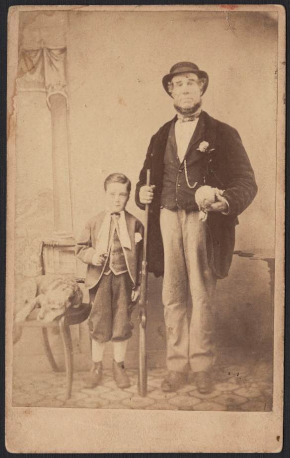 Photo noir et blanc montrant un homme, fusil à la main, debout aux côtés d’un garçon; près d’eux, un chien est couché sur une chaise.