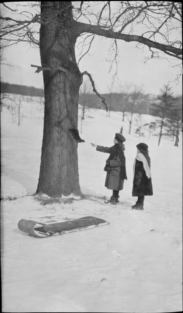 Photographie noir et blanc de deux jeunes filles debout près d’un arbre, en hiver. Une des jeunes filles nourrit un écureuil accroché au tronc.