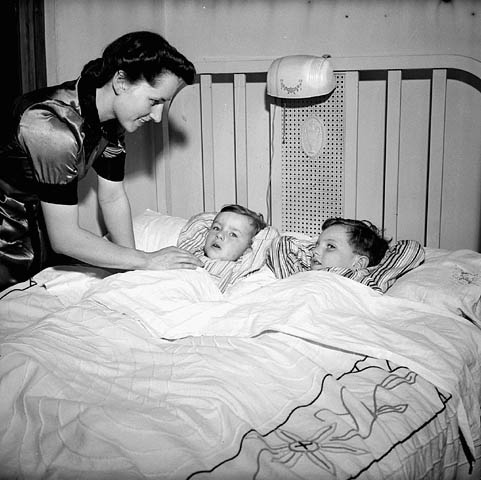 Photo noir et blanc d’une mère bordant ses deux fils. 
