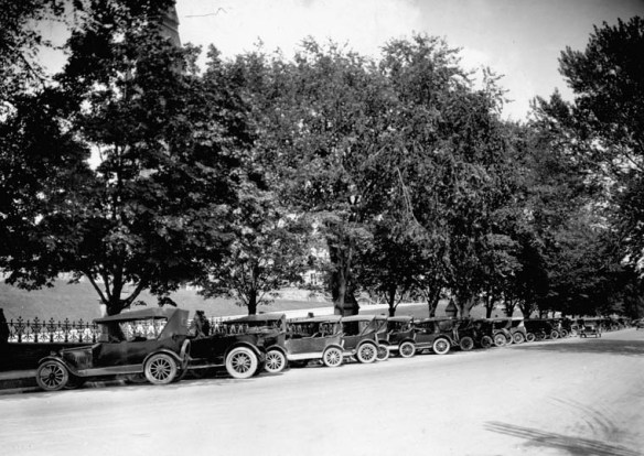 Photographie noir et blanc d’une rue bordée d’arbres avec une longue rangée d’autos stationnées de biais le long du trottoir.
