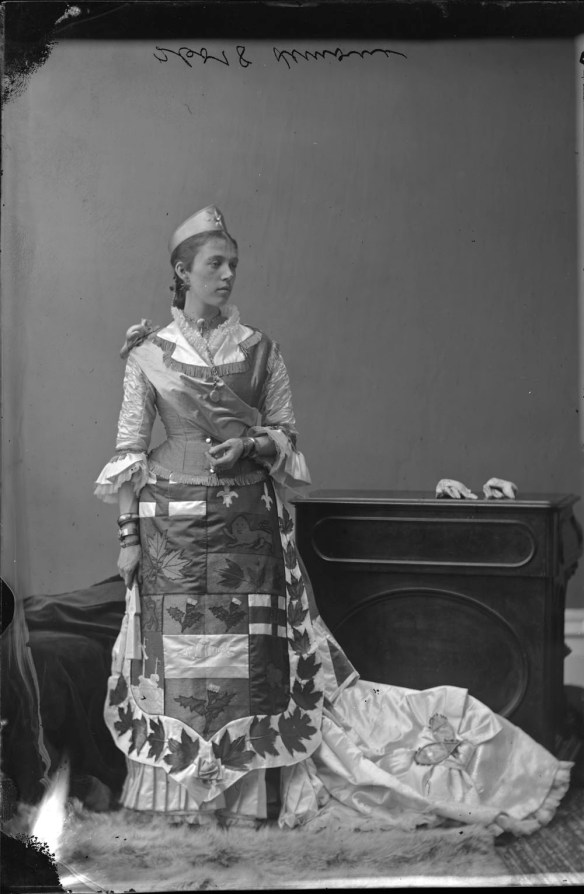 Photo noir et blanc d’une femme costumée en Dominion du Canada dans un studio de photographie. Sur le devant de sa robe, on aperçoit les armes du Dominion entourées de feuilles d’érable brodées. Des raquettes miniatures se trouvent sur la traîne de sa robe.
